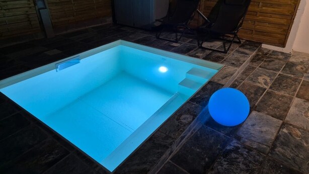 Una piscina rectangular de tono azul claro, rodeada de baldosas de piedra oscura. Se observa una silla negra y un calentador al fondo.