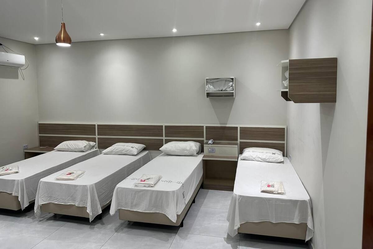 Une chambre d’hôtel avec quatre lits simples, une tête de lit en bois, une petite table de chevet, un climatiseur fixé au mur et des spots encastrés au plafond.