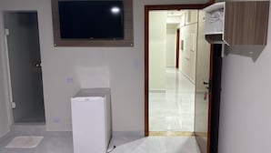 Una habitación con un televisor de pantalla plana montado en la pared, un refrigerador blanco pequeño y un armario de madera con un perchero.