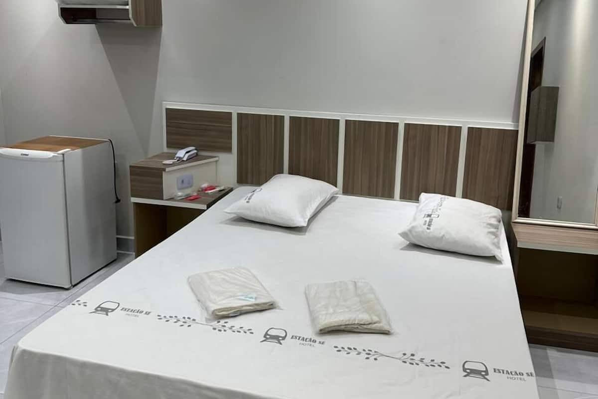 Une chambre d’hôtel moderne avec un lit bien fait, une petite table de chevet, un miroir et un réfrigérateur compact.