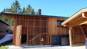 A modern wooden building with a large open entrance, a staircase leading to an upper level, and a stone wall adjacent to the entrance.