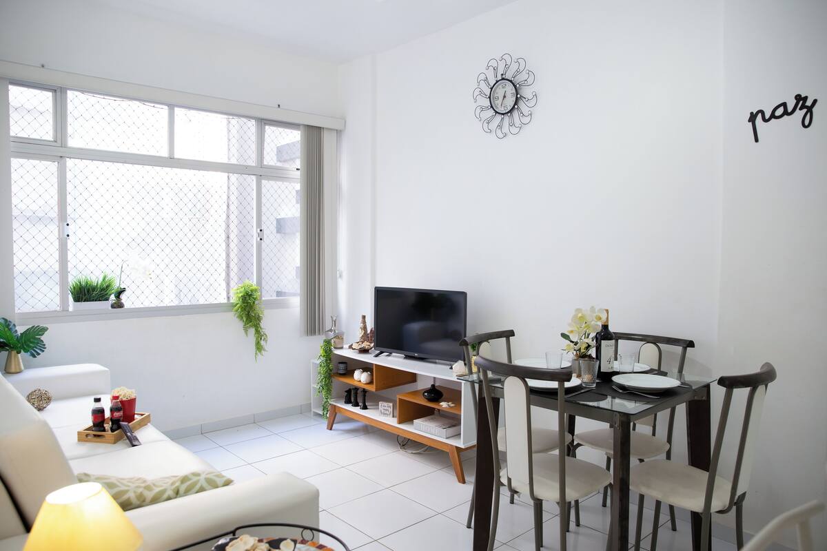 A modern living room with a sofa, TV, dining table, and chairs. There is a window with plants and a decorative wall piece.