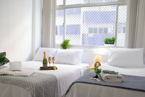 Une chambre d’hôtel avec deux lits, une grande fenêtre avec des rideaux, une plante en pot, une bouteille de champagne et une lampe.