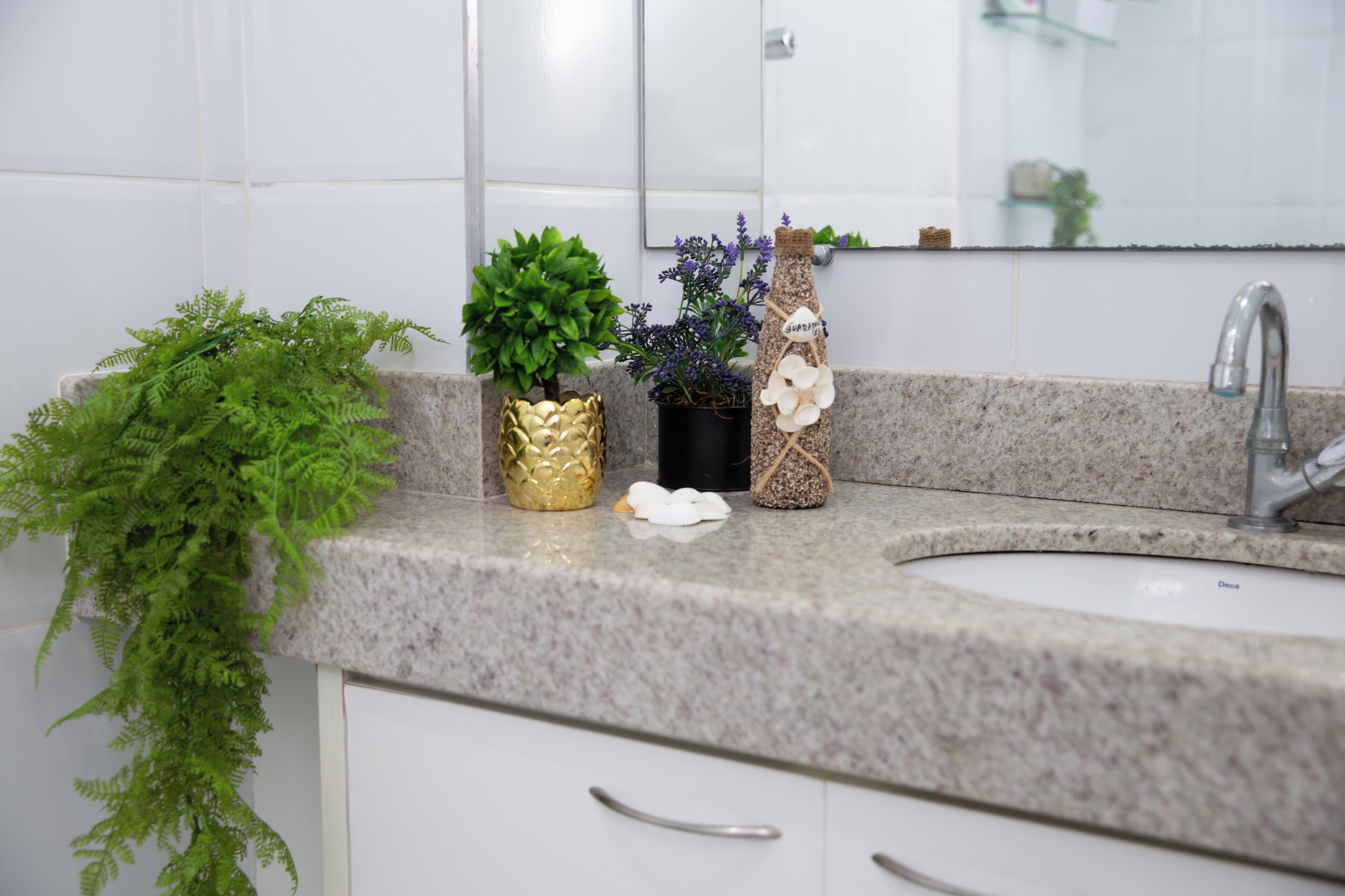 A bathroom with a granite countertop, a white sink, and a mirror above it. There are three potted plants on the countertop: one with green leaves, one with purple flowers, and one with white flowers.