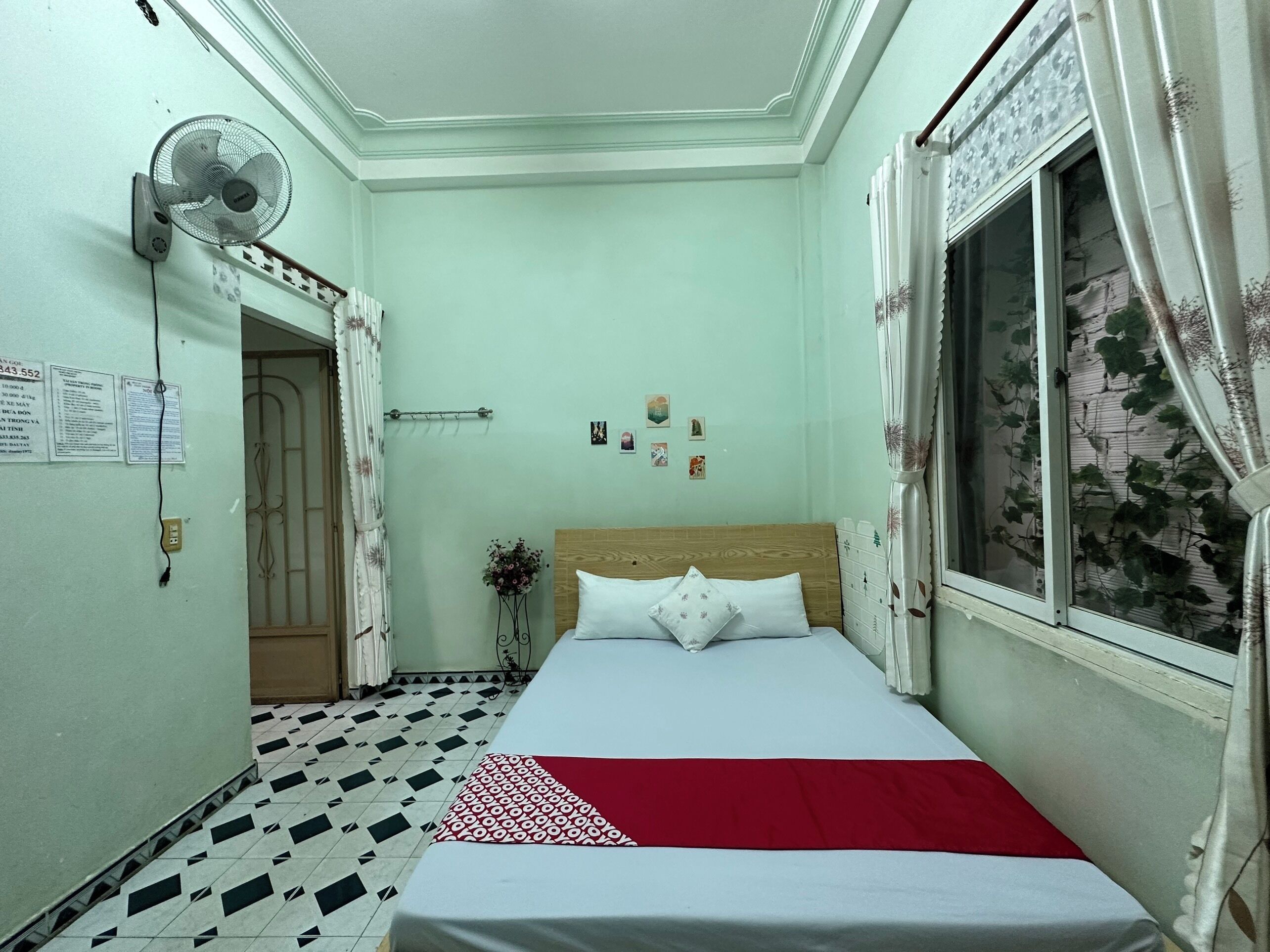 A narrow hotel room with a bed, a standing fan, a window with curtains, and a tiled floor.