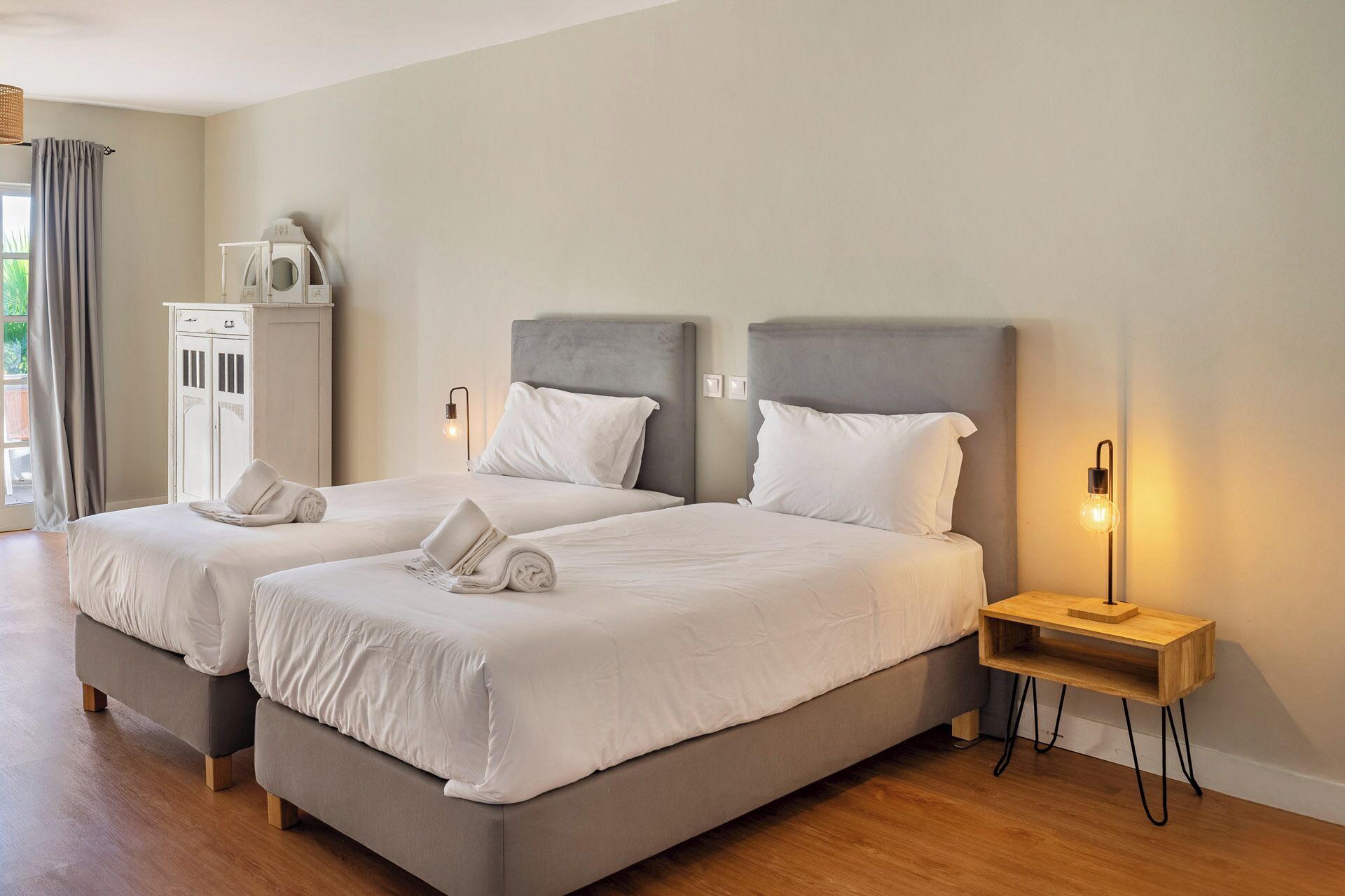 Habitación doble con dos camas, cada una con cabeceros grises y ropa de cama blanca. Un mesita de noche de madera con lámpara a un lado, y un armario con espejo al otro.
