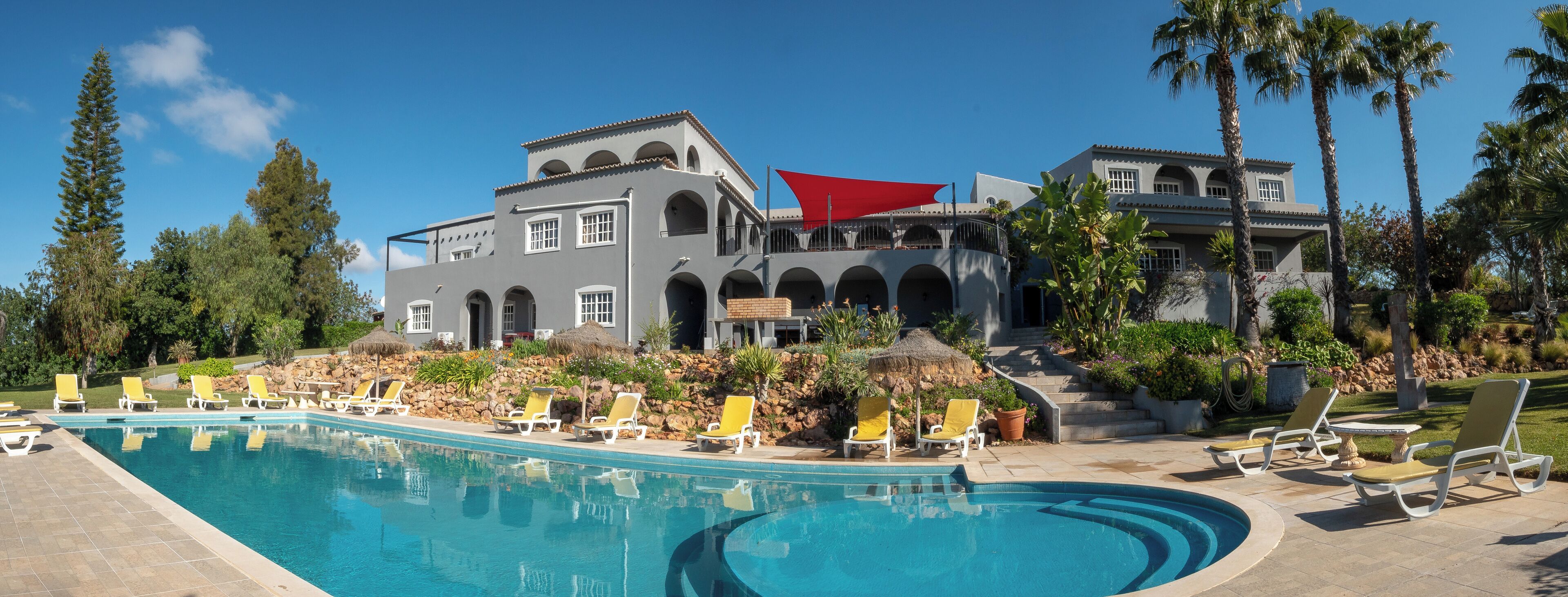 Una piscina grande con tumbonas, un toldo rojo y un edificio con ventanas de arco y un muro de piedra.