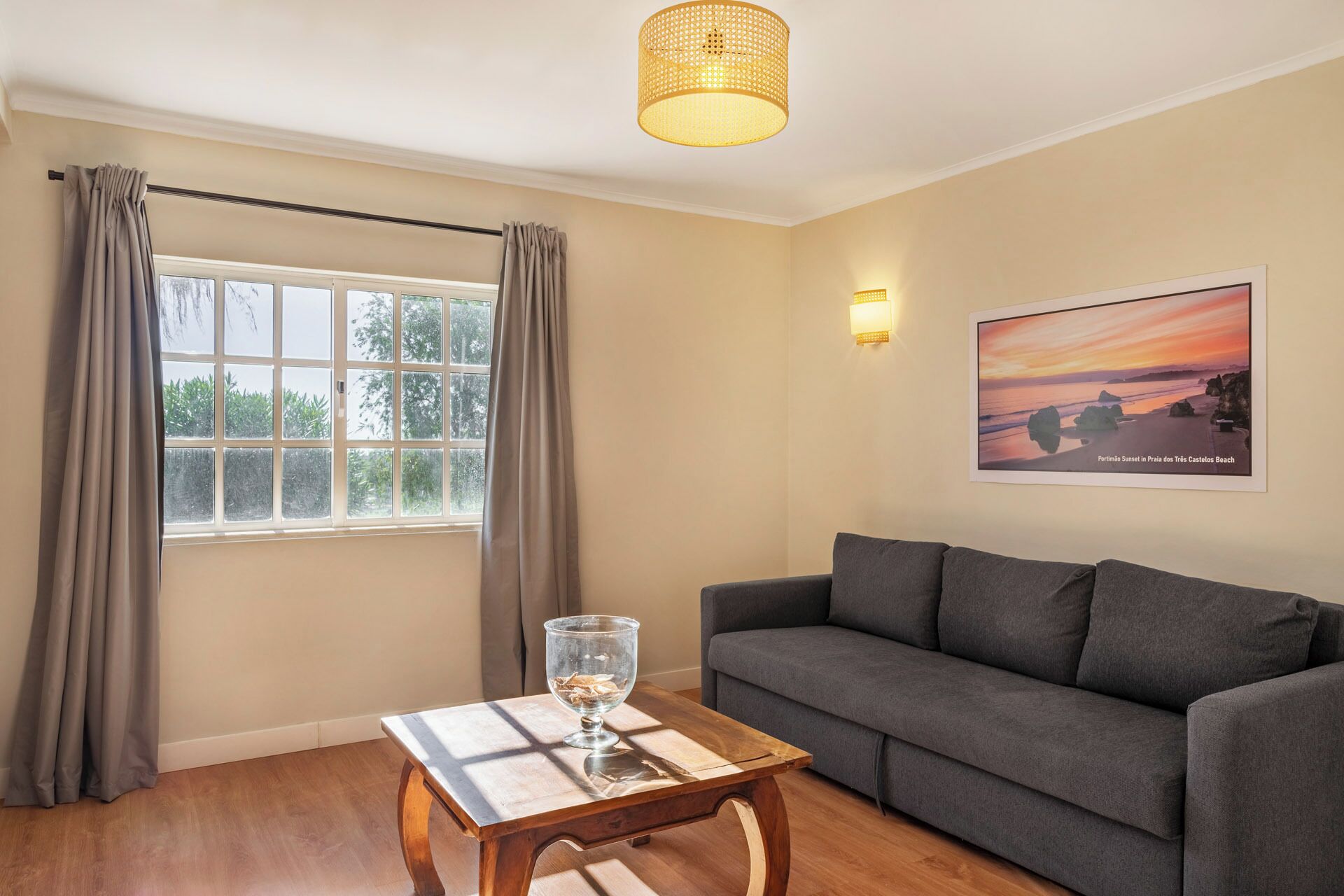 Una sala de estar con un sofá gris, una mesa de centro de madera con un florero de vidrio, una ventana con cortinas, una lámpara de pared y un cuadro enmarcado con una playa al atardecer.