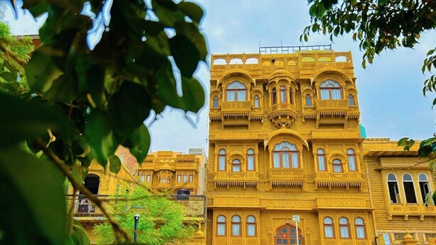 A large, ornate building with multiple windows and balconies, surrounded by greenery.