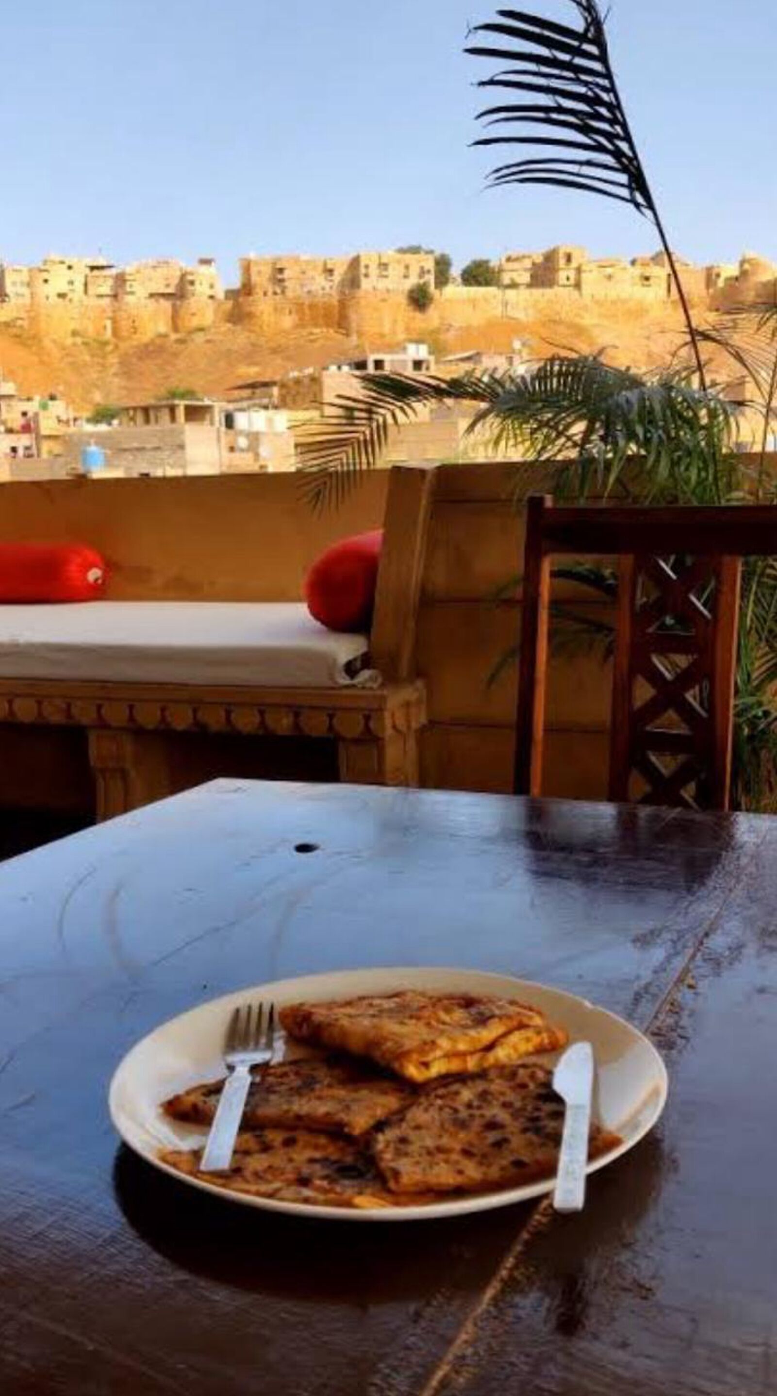 Una mesa de comedor con un plato de comida y cubiertos, una silla, y vista a un paisaje urbano con edificios al fondo.
