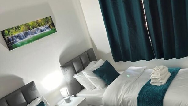 Hotellihuone, jossa on kaksi sänkyä. Jokaisessa sängyssä on valkoinen peitto ja turkoosit yksityiskohdat. Huoneessa on yöpöytä, jossa on lamppu, ja seinällä on taulu, jossa on kuva vesiputouksesta.