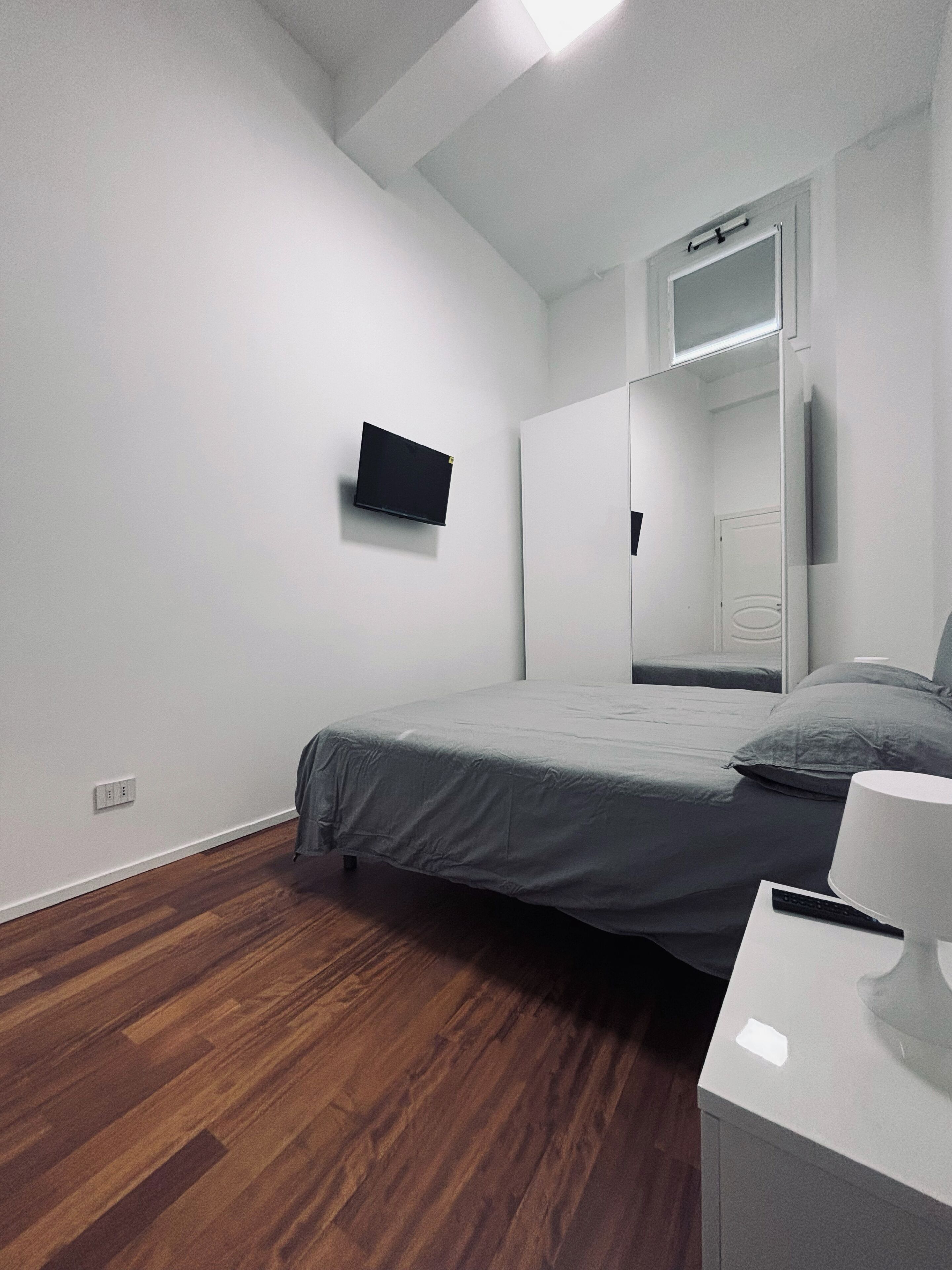A bedroom with a bed, a bedside table with a lamp, a wall-mounted TV, and a mirror.