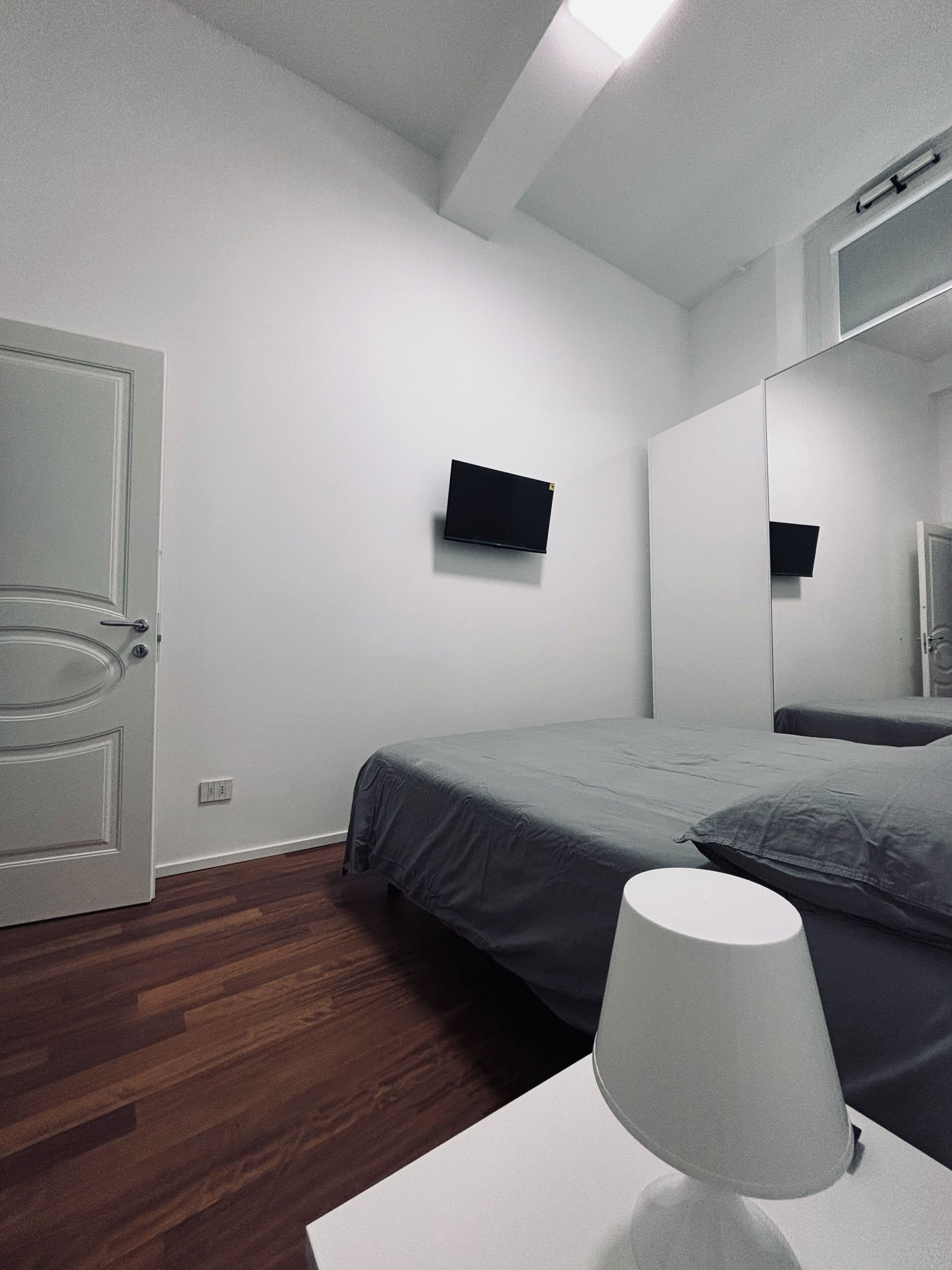 Un dormitorio con cama, mesita de noche con lámpara, televisor montado en la pared y una puerta.