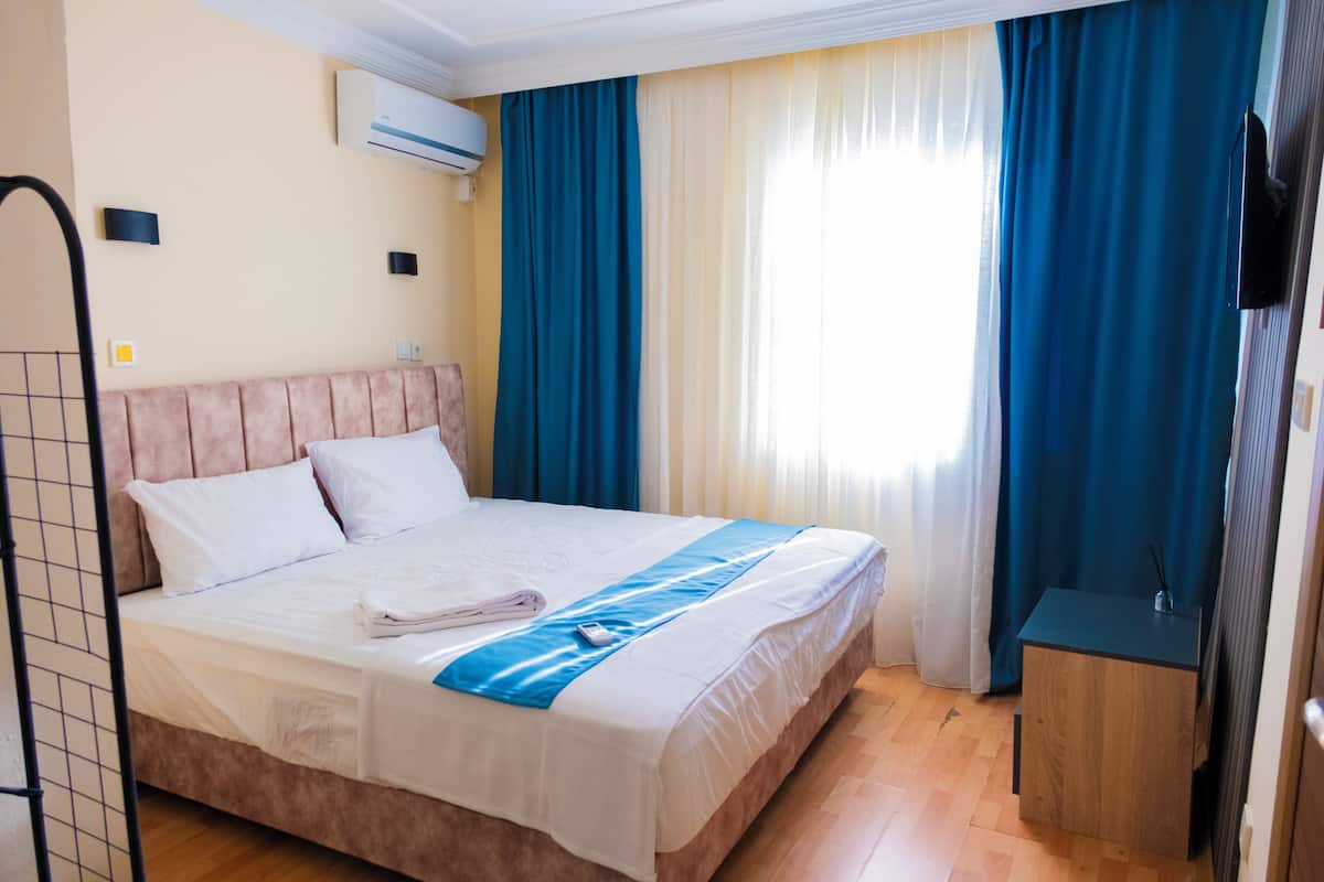 A hotel room with a bed, a wooden nightstand, a wall-mounted TV, and a window with curtains.