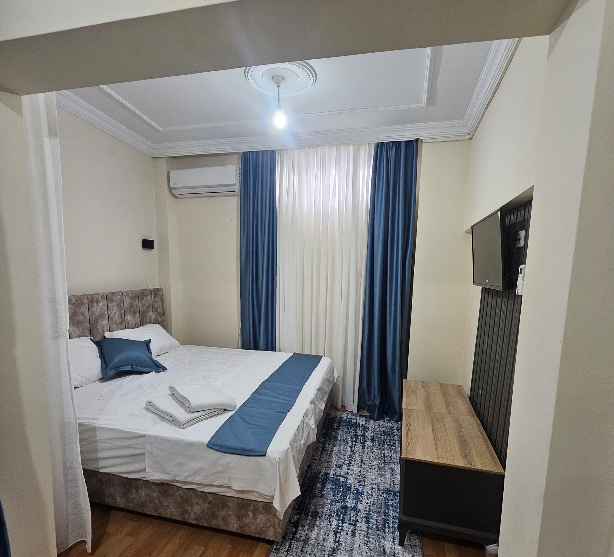 Habitación Galaxy S23 Ultra con una cama, unidad de aire acondicionado, cortinas, televisor y una mesita de noche de madera.