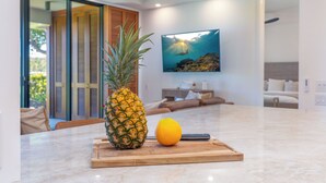 Une cuisine avec un ananas et une orange sur une planche à découper, une table à manger et un coin salon avec un canapé et un tableau représentant une scène sous-marine.