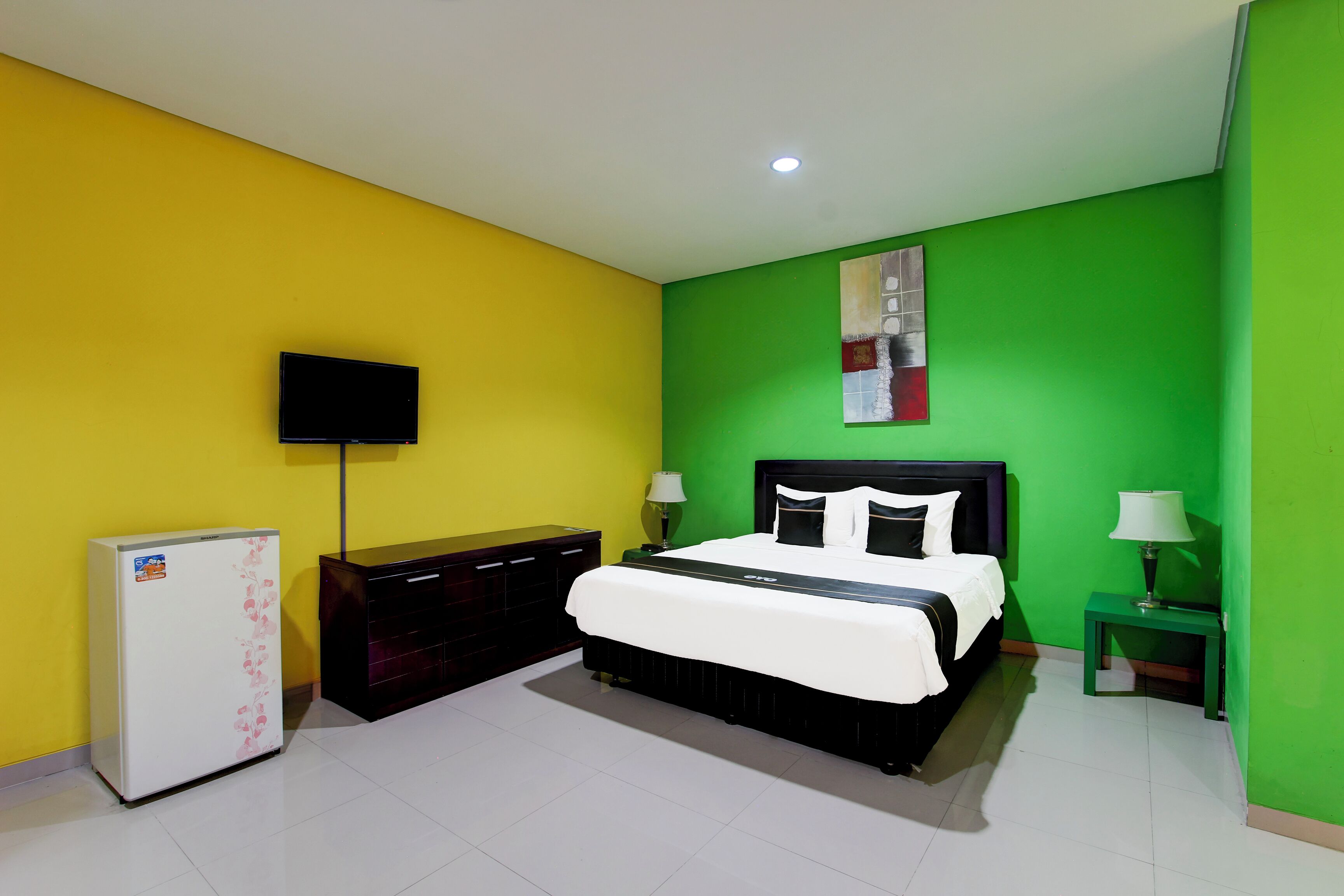 Un dormitorio con cama, televisión, refrigerador y mesita de noche con lámpara.