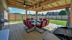 Een overdekt terras met een houten tafel en stoelen, een barbecue en een uitzicht op een huis op de achtergrond.