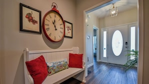Een gang met een witte bank, rode kussens, een grote wandklok, ingelijste kunst en een deur naar een andere kamer.
