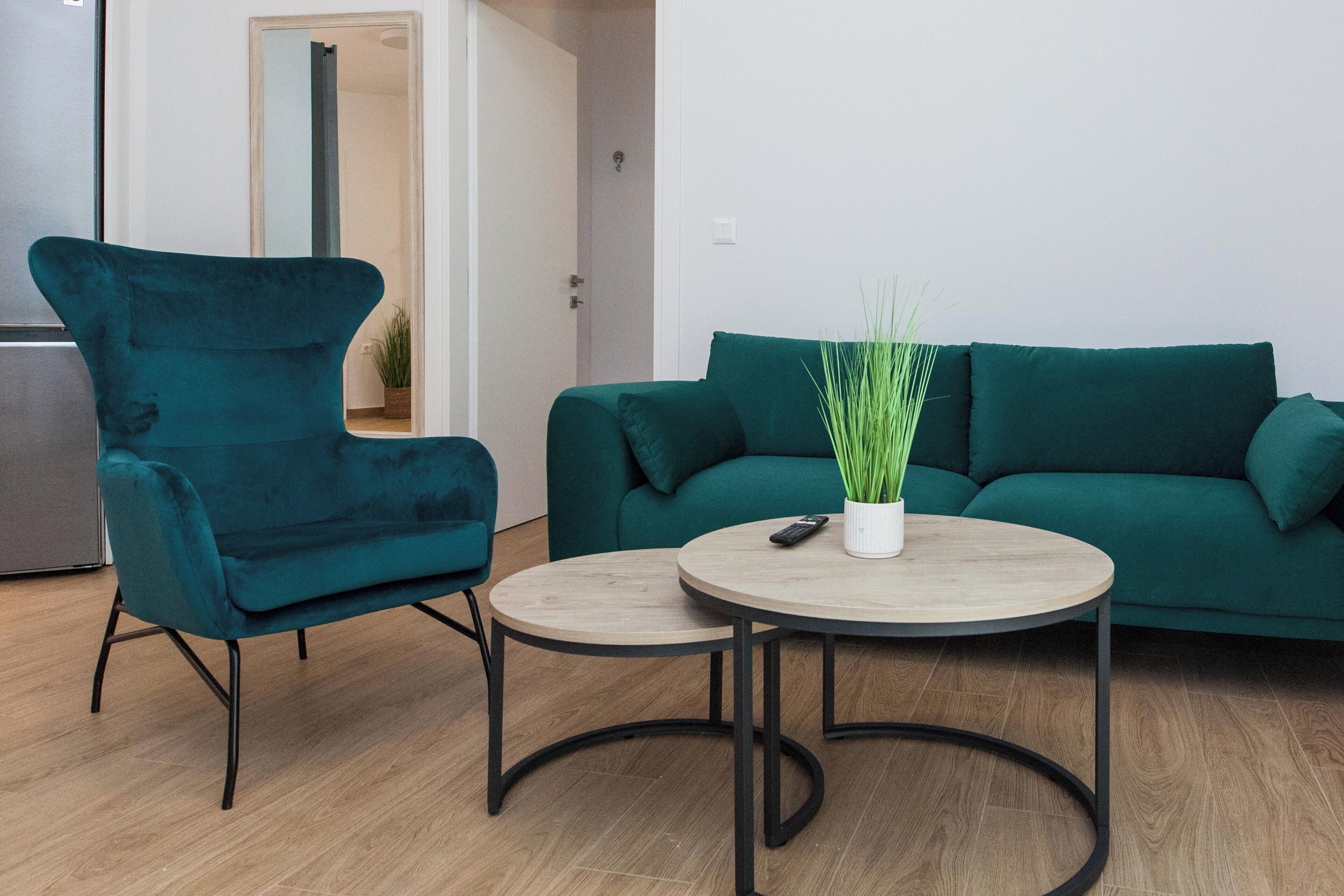 Een moderne woonkamer met een blauwgroene fauteuil, een groene bank en een ronde houten salontafel met een afstandsbediening en een potplant.