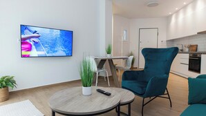 Egy modern nappali, melyben egy síkképernyős televízió, egy kerek, fából készült asztal, egy türkiz színű karosszék és a háttérben egy konyhasarok található.