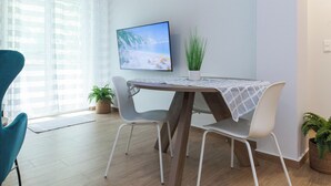 Egy modern étkező, melyben egy fa asztal, fehér székek és a falra szerelt televízió található.