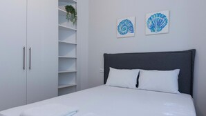 Egy gondosan megterített ágy fehér ágyneművel és párnákkal, szürke ágyvégfal, és két, keretbe foglalt műalkotás a falon.