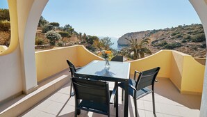 En balkon med et bord og stole, der giver udsigt over et kystlandskab med klipper og vegetation.