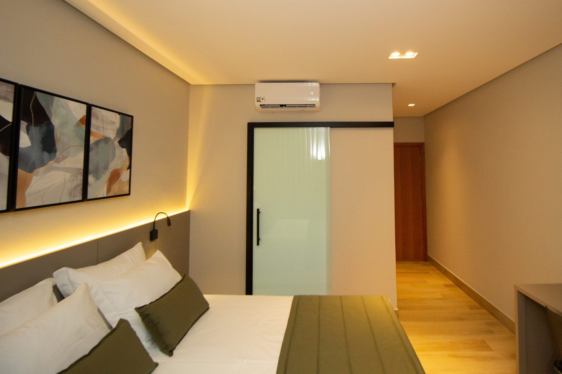 Habitación de hotel con cama, mesita de noche, aire acondicionado instalado en la pared y puerta que da a un pasillo.