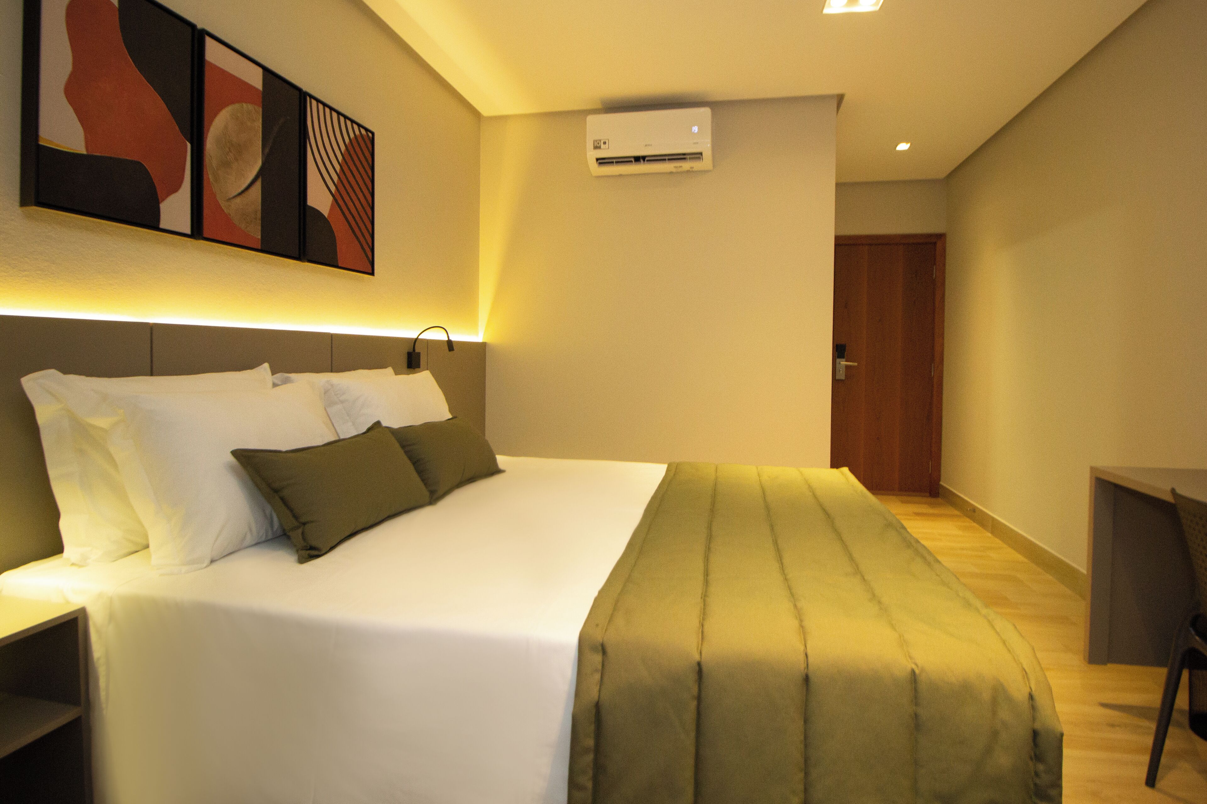 A modern hotel room with a neatly made bed, a bedside table, a desk, and an air conditioning unit on the wall.