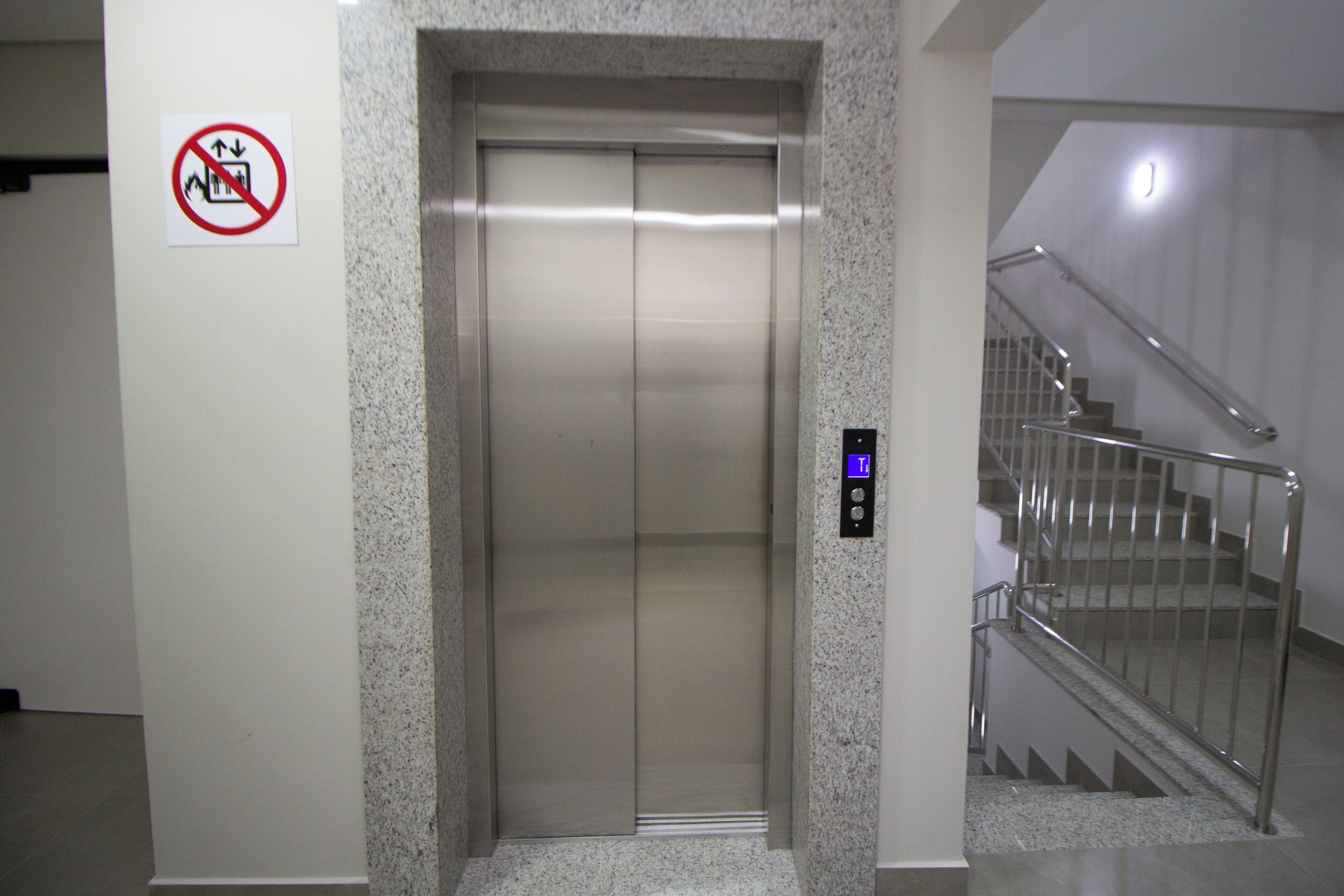 Um elevador com placa de proibido fumar, painel de comando e escada com grades de metal.