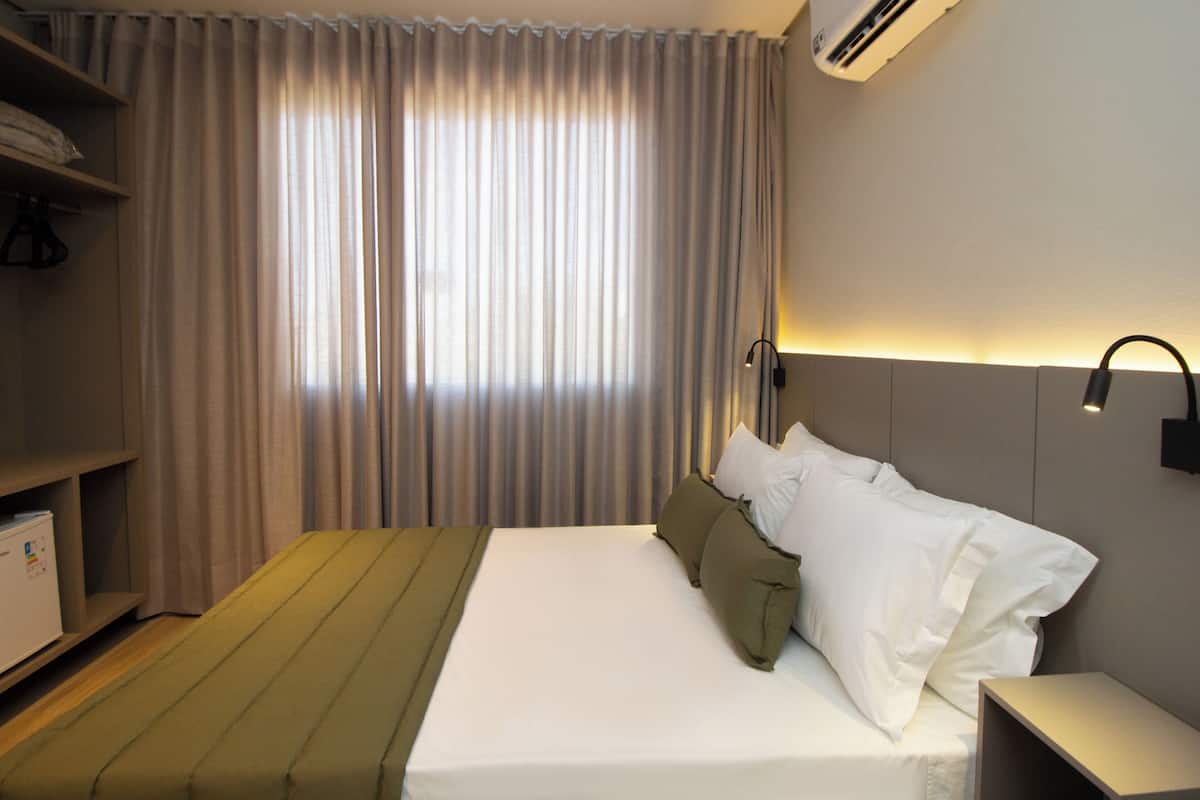 A hotel room with a bed, a bedside table, a shelf with a small appliance, and a window with curtains.