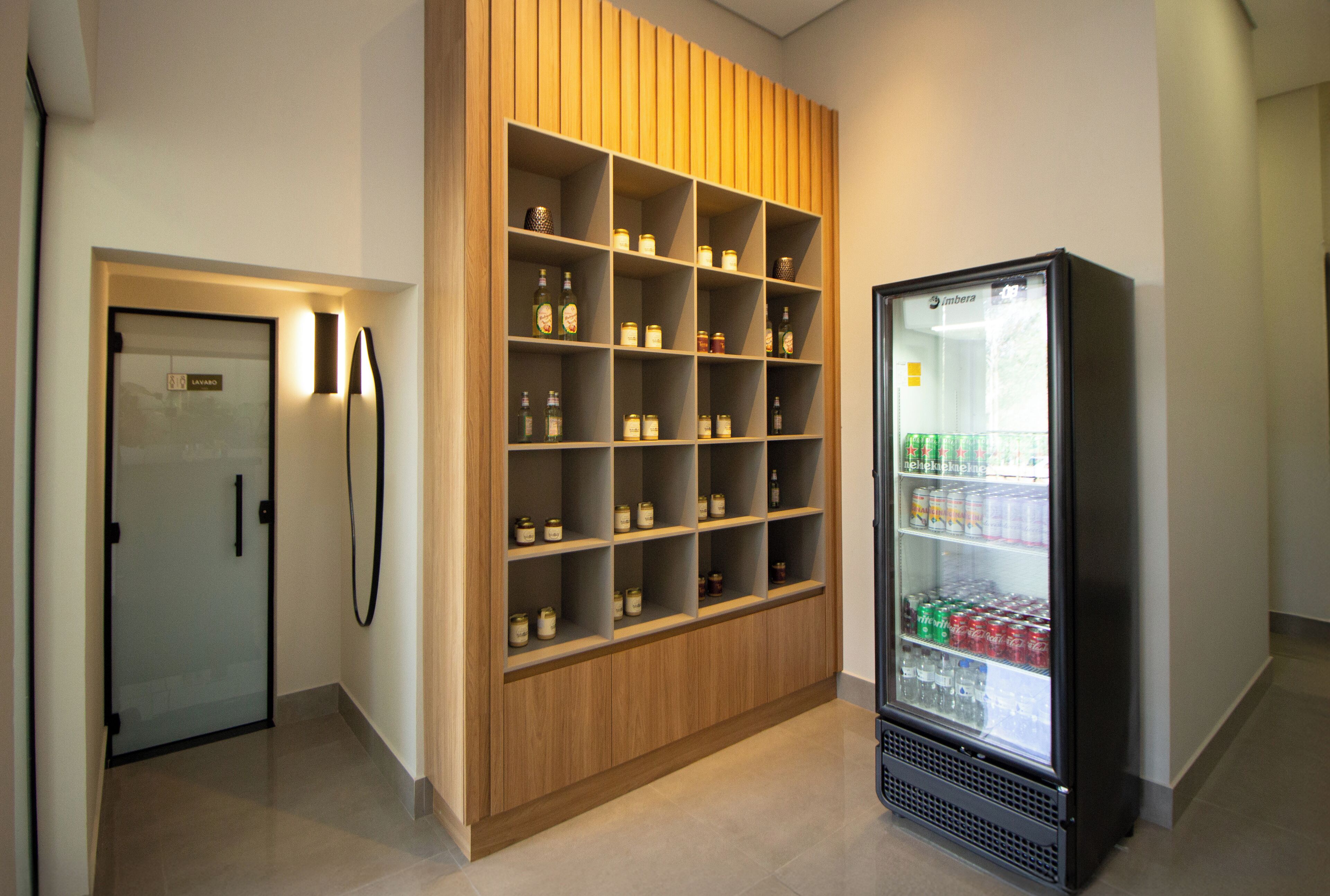 A modern hotel room with a wooden shelving unit filled with jars and a minibar refrigerator stocked with beverages.
