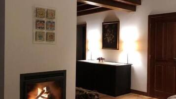 A room with a fireplace, wooden flooring, a high ceiling with exposed beams, a small table with a vase, and a door.