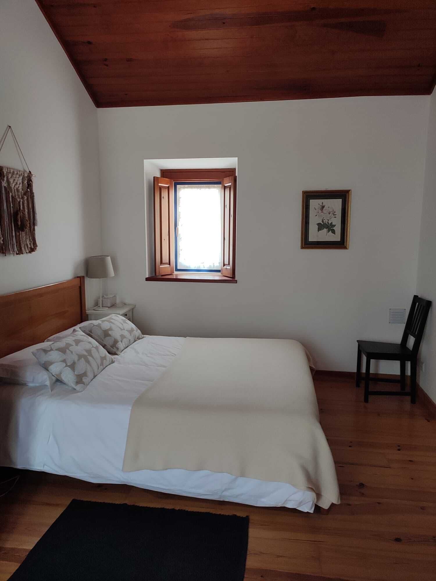 A bedroom with a wooden ceiling, a bed with white bedding, a window with wooden shutters, a framed picture on the wall, and a chair.