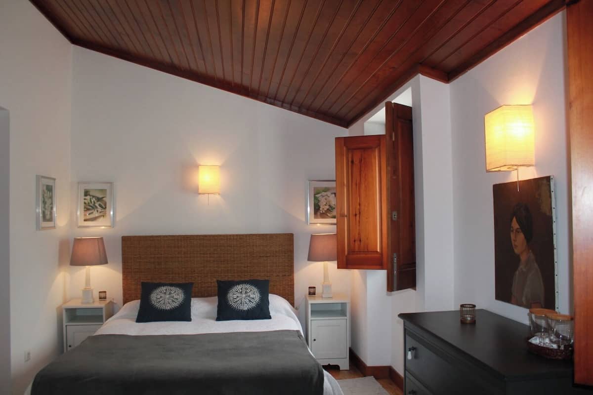 A bedroom with a wooden ceiling, two bedside tables with lamps, a bed with a dark headboard, and a dresser with a mirror.