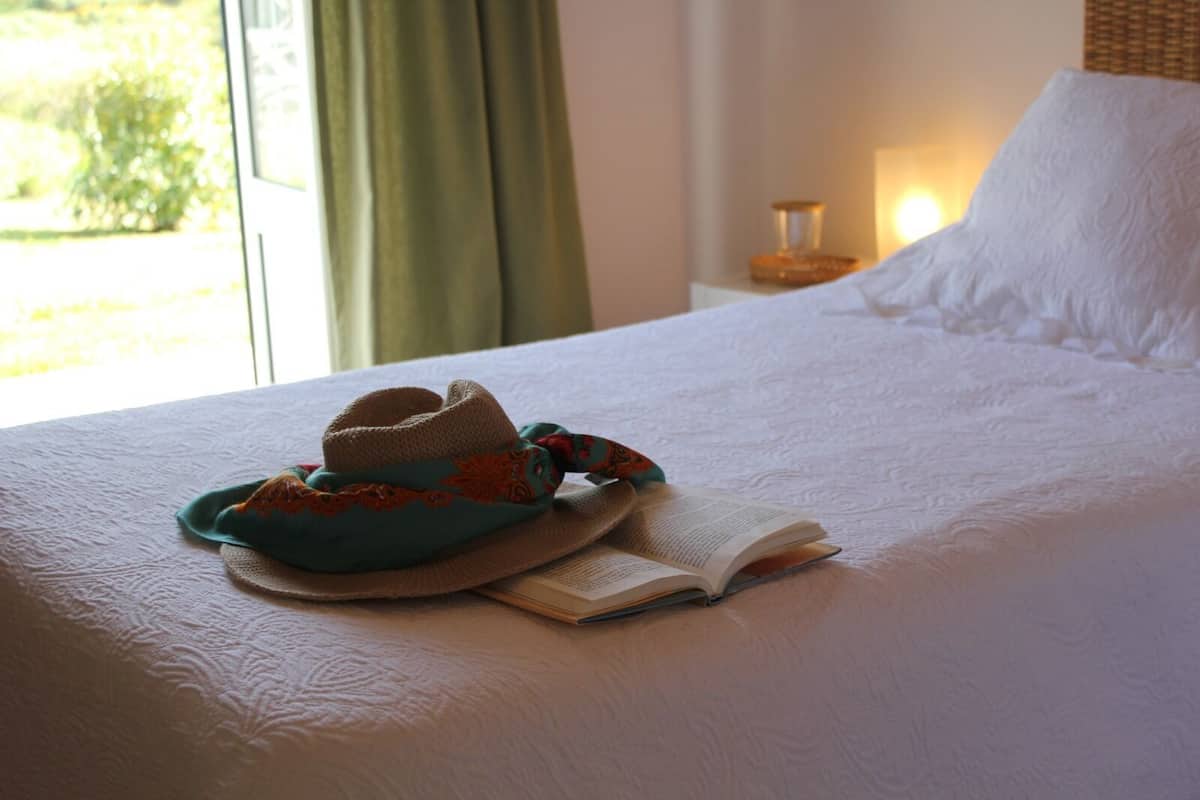 A neatly made bed with a hat and book on it, a bedside table with a lamp, and a window with curtains.