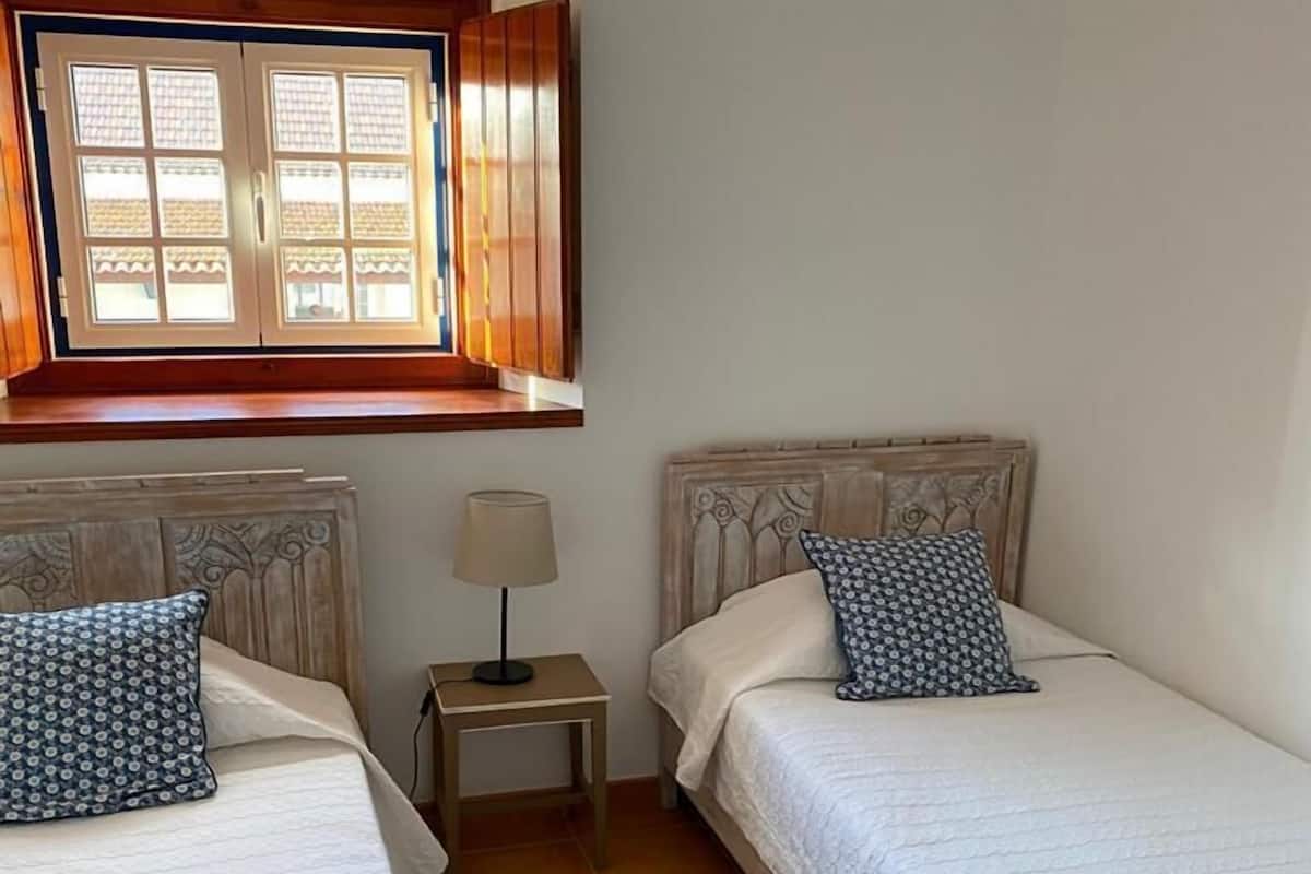A small, cozy bedroom with two single beds, a nightstand with a lamp, and a window with wooden shutters.
