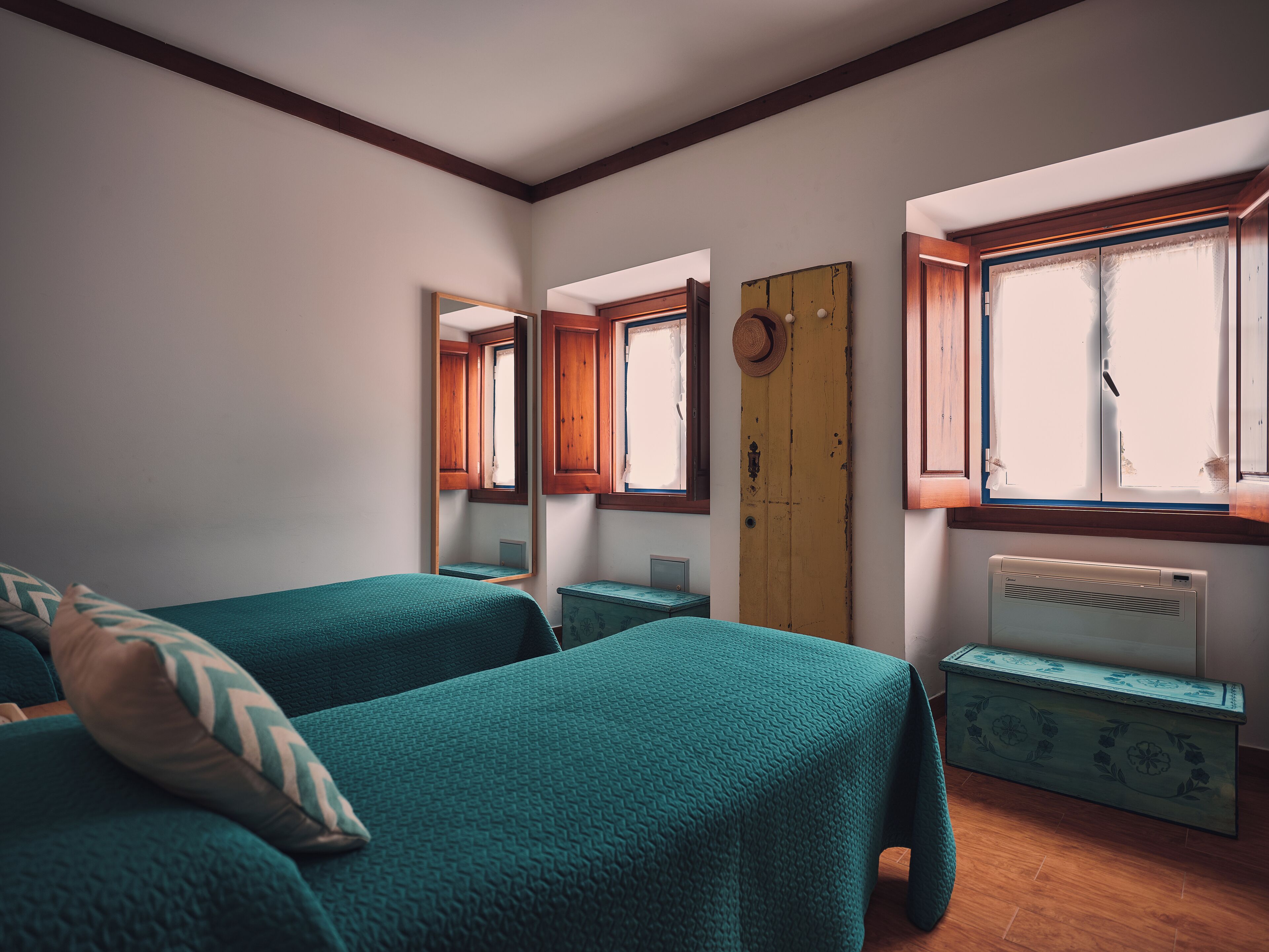 Quarto com duas camas, porta de madeira, radiador e janela com cortinas.