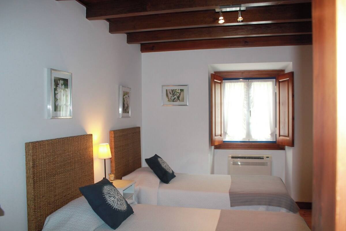 A hotel room with two beds, a window with curtains, a wall-mounted air conditioner, and framed pictures on the wall.