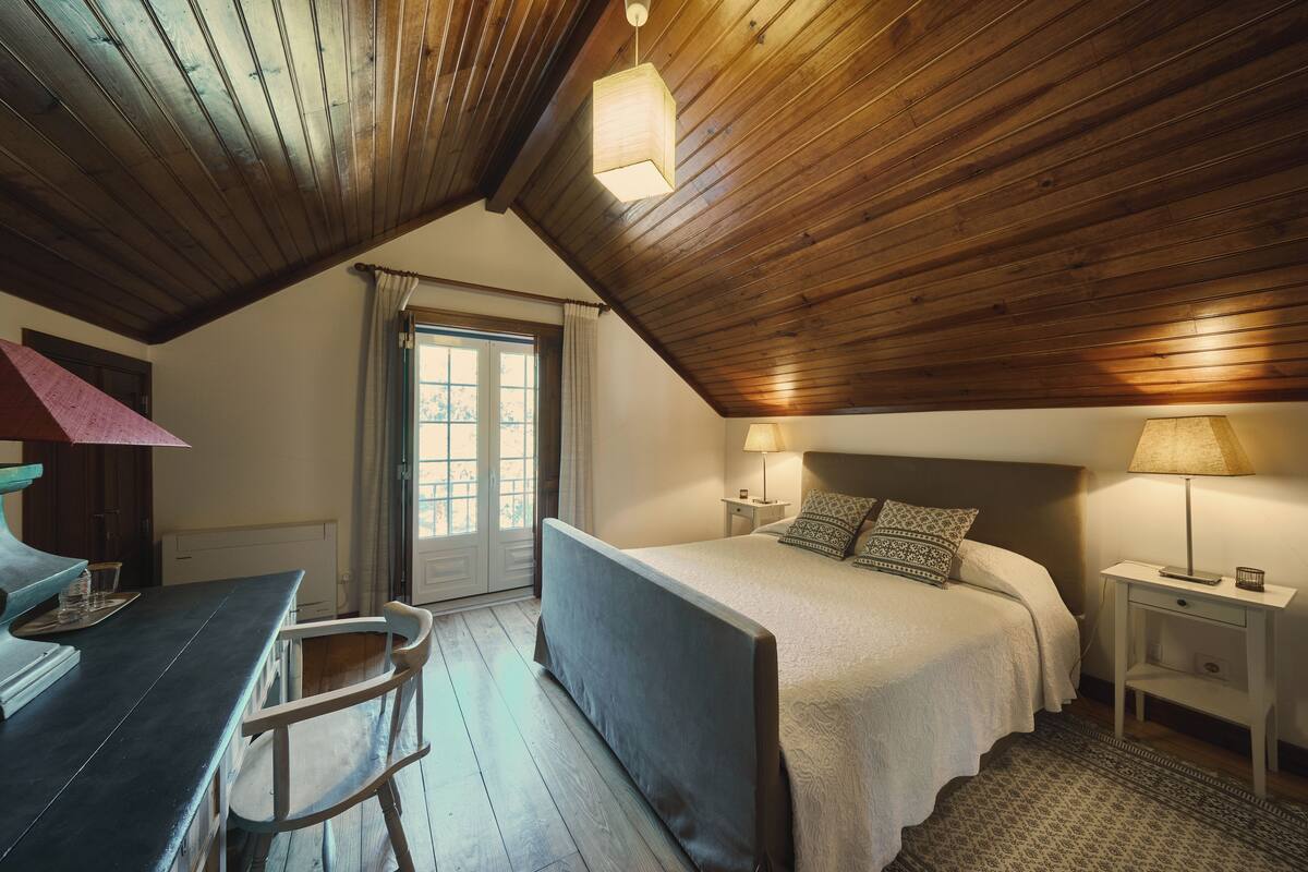 A bedroom with a wooden ceiling, a bed with a patterned bedspread, two bedside lamps, a nightstand, and a window with curtains.