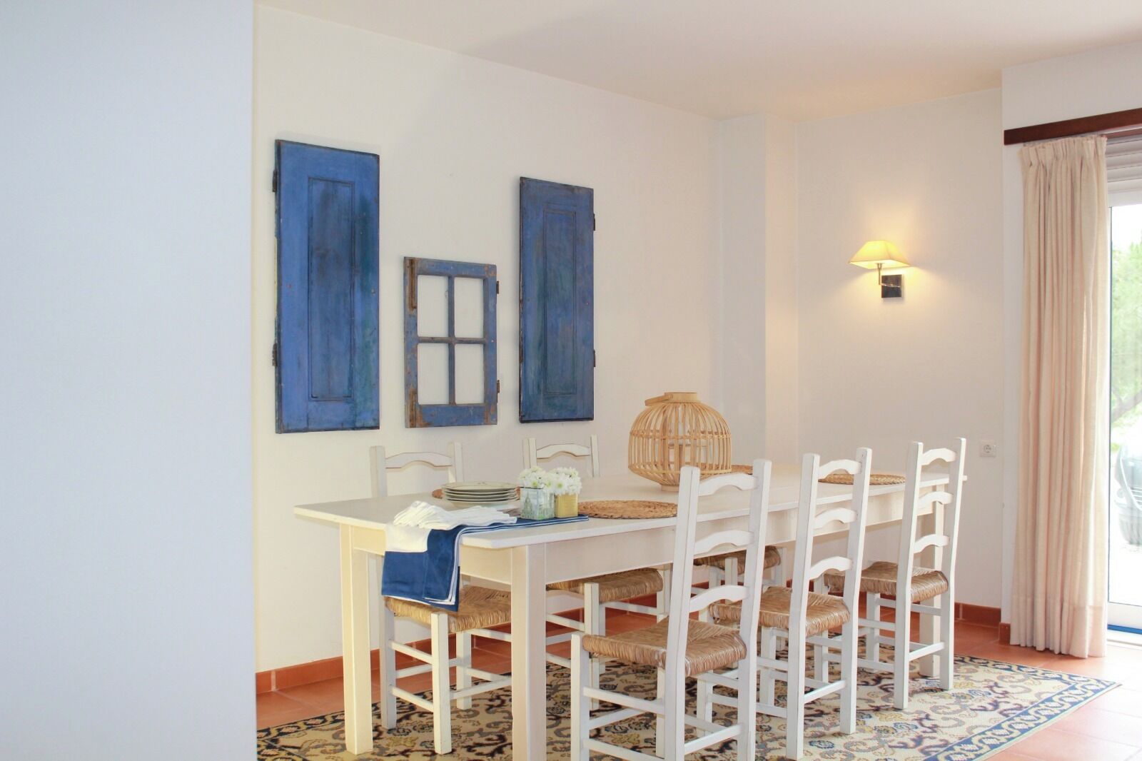 A dining area with a white table and chairs, blue shutters on the wall, a woven basket, and a lamp.
