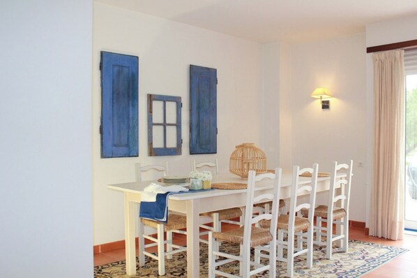 Un comedor con una mesa y sillas blancas, contraventanas azules en la pared, una cesta tejida y una lámpara.