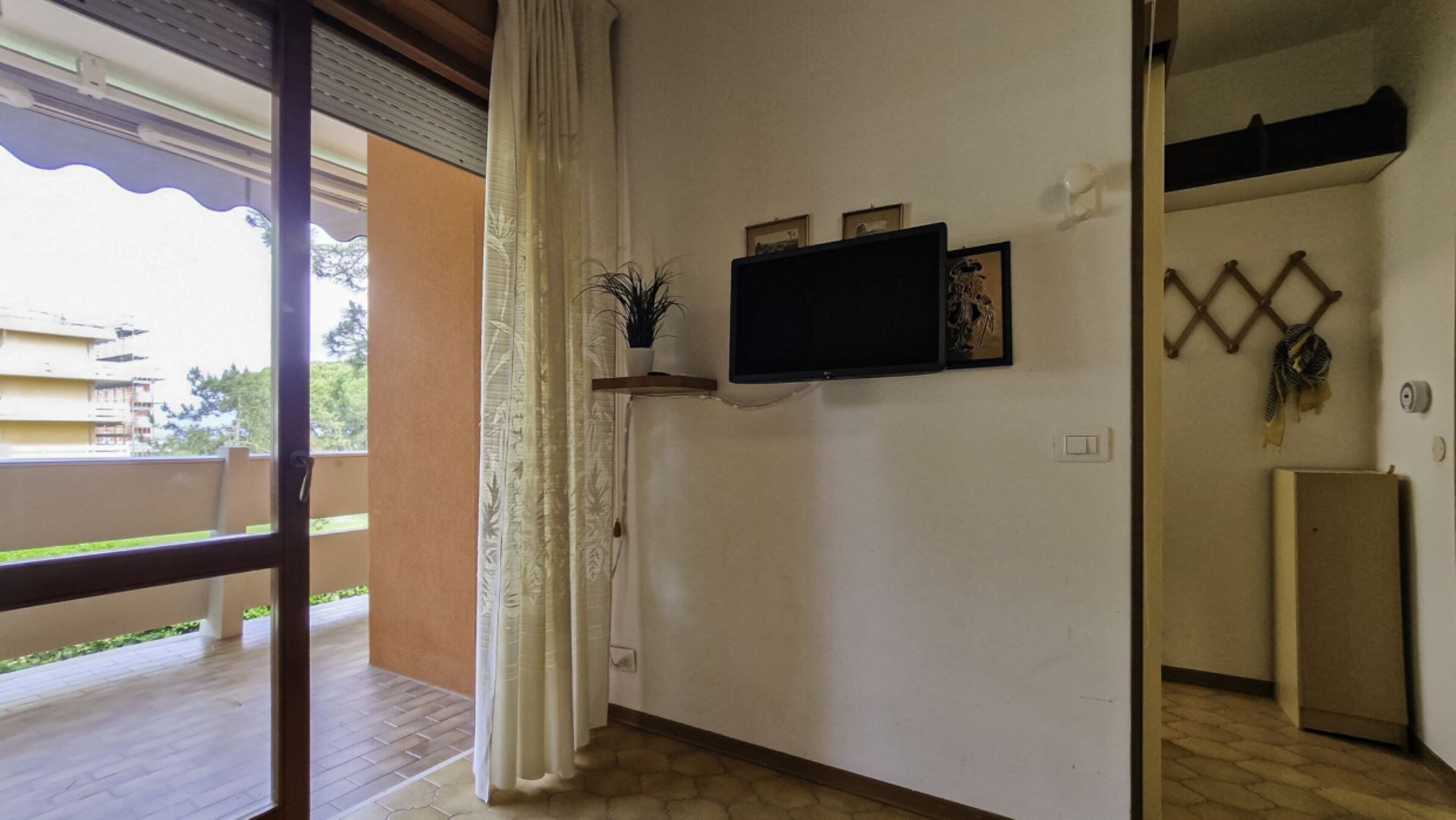 A room with a TV mounted on the wall, a small shelf with decorative items, a window with curtains, and a view of the outside balcony.