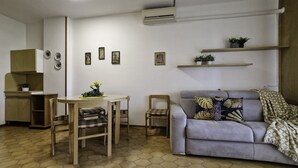 En stue med sofa, spisebord med stole, et køkkenområde med skabe og vægmonterede hylder med planter.