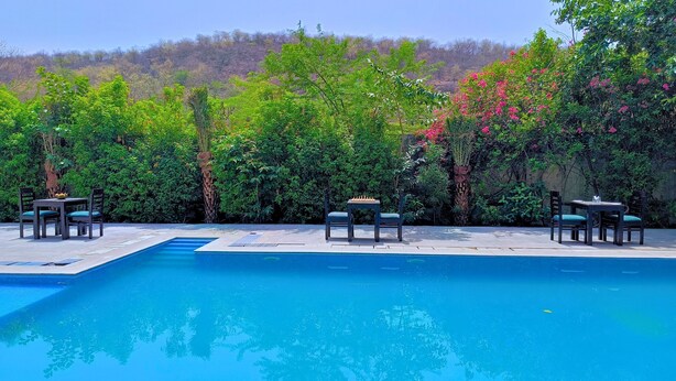 A swimming pool with four tables and chairs, surrounded by lush greenery and flowering plants. - Maharana Palace by Asapian Hotels (Amer)