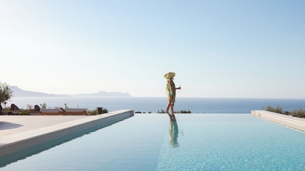 A person standing at the edge of a infinity pool with a view of the sea and mountains in the background.