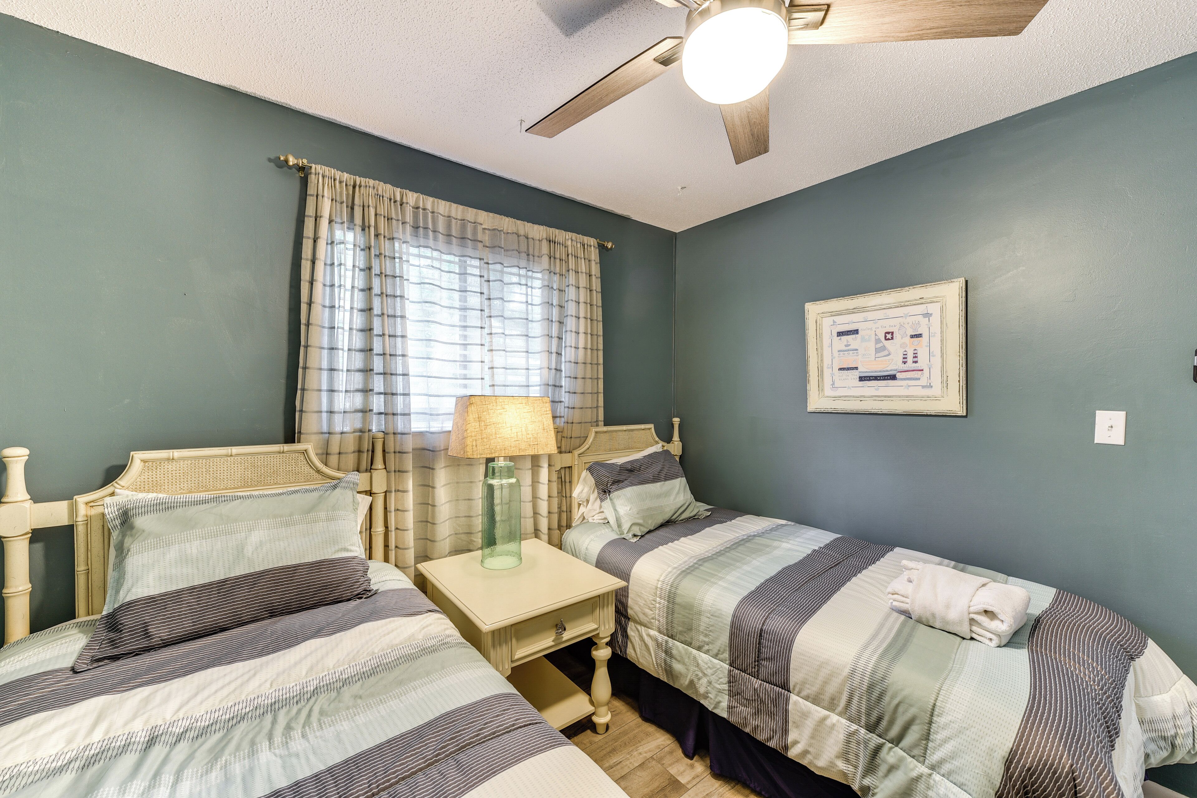 A room with two beds, a ceiling fan, a window with curtains, and a wall-mounted light switch.