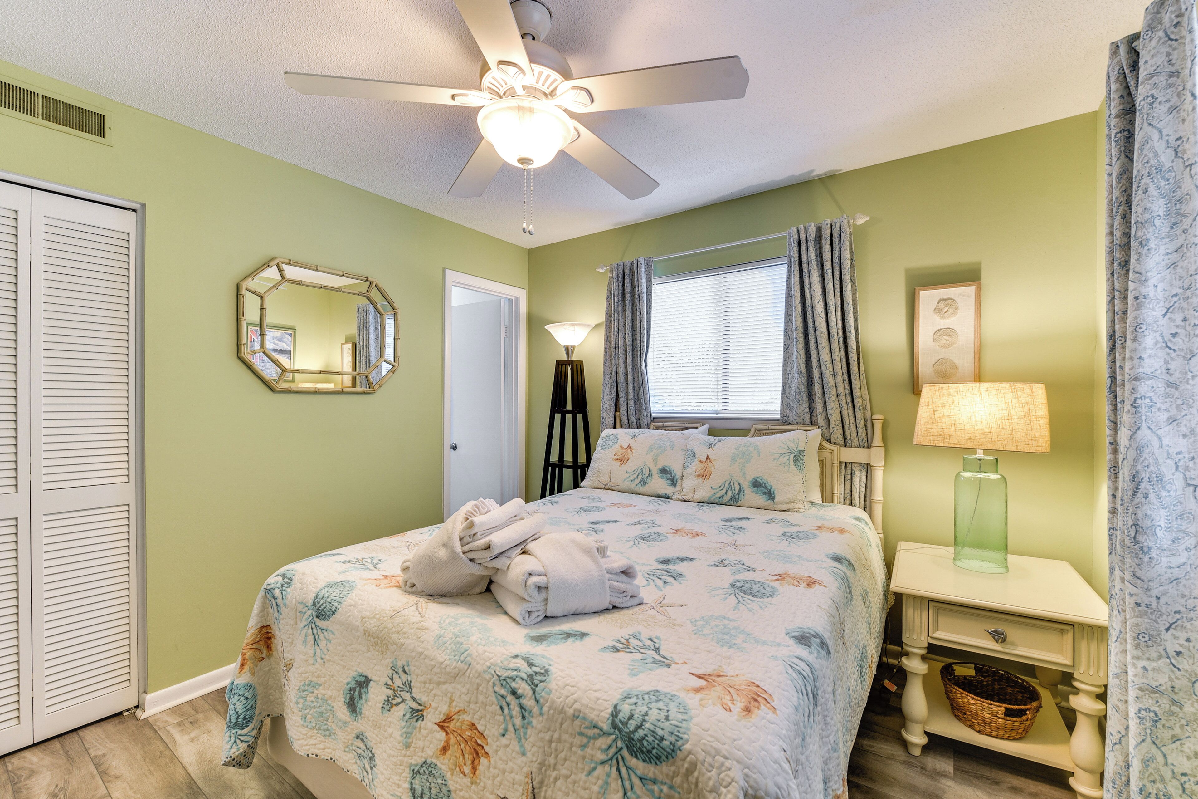 A bedroom with a bed, a ceiling fan, a nightstand, a lamp, a mirror, and a window with curtains.