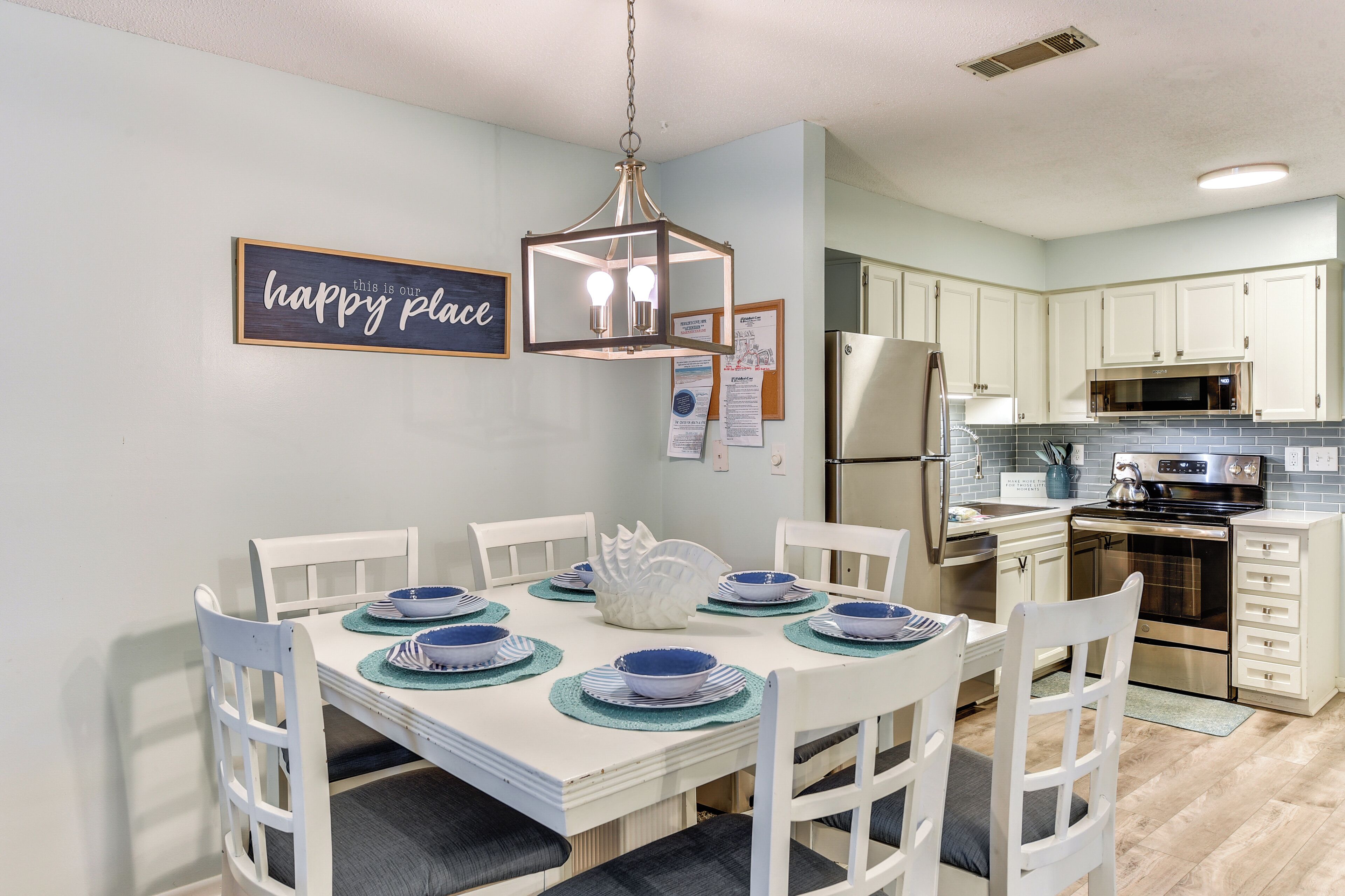 A dining area with a white table set for six, surrounded by white chairs. A chandelier hangs above, and a 'happy place' sign is on the wall. The kitchen in the background has white cabinets and stainless steel appliances.